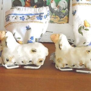 Puede incluir: Dos figuras de ovejas de cerámica blanca con manchas marrones y bases de metal. Las ovejas tienen un aspecto rústico y envejecido. Detrás, tela estampada con flores y una imagen enmarcada de gallinas.