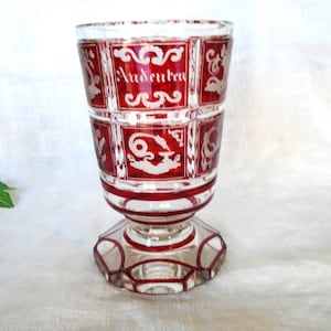 May include: A clear glass goblet with a red, geometric design. The goblet features the word "Audentes" in white lettering. The base is a clear, multi-sided shape. The goblet is likely used for drinking.