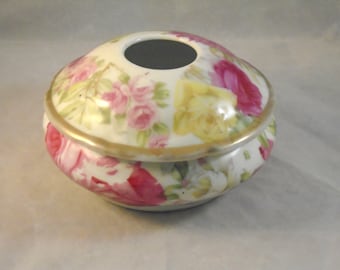 May include: A white porcelain trinket box with a lid. The box is decorated with a floral pattern of pink, yellow, and red roses. The lid has a gold trim and a small hole in the center.