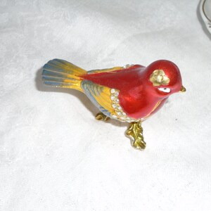 May include: A red, yellow, and blue bird figurine with gold accents and rhinestones. The bird is perched on a gold stand with a leaf design.