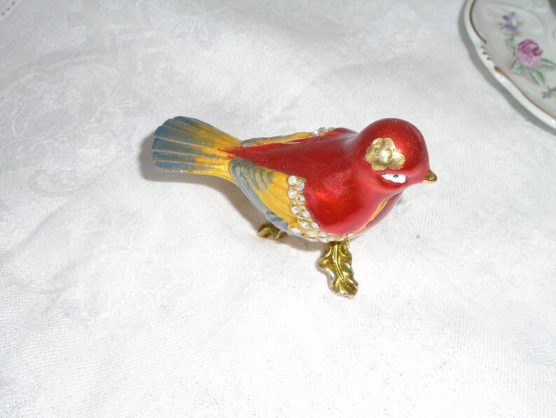 May include: A red, yellow, and blue bird figurine with gold accents and rhinestones. The bird is perched on a gold stand with a leaf design.