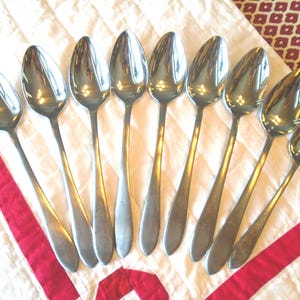 May include: A set of nine silver-toned metal spoons arranged in a fan-like pattern. Each spoon features a smooth, rounded bowl and a plain, slender handle. The spoons have a polished, reflective finish and are uniform in their simple, functional design.