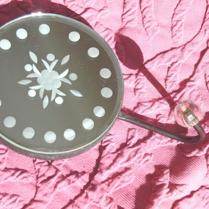 May include: A round, decorative mirror with a floral design etched in the center and white dots around the edge. The mirror is attached to a black metal hook with a clear, round glass knob. The mirror rests on a pink, quilted surface.