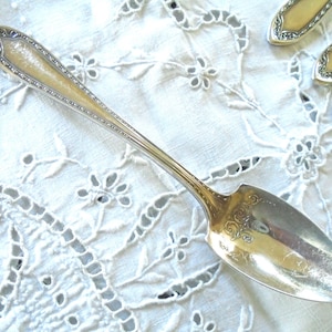 Könnte beinhalten: Ein silberner Löffel mit einem verzierten Griff und einem dekorativen Design auf der Schüssel. Der Löffel liegt auf einem weißen, bestickten Stoff, im Hintergrund sind weitere Besteckteile zu sehen.