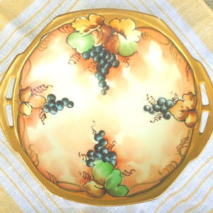 Puede incluir: Una bandeja de servicio redonda de cerámica con dos asas laterales, que presenta un diseño pintado a mano de racimos de uvas azul oscuro y hojas verdes y naranjas sobre un fondo crema y melocotón, con un borde de color dorado.
