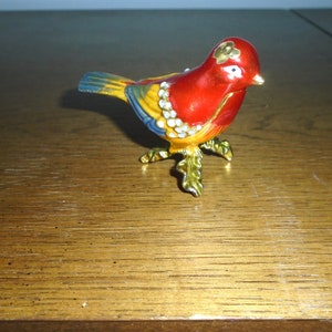 May include: A small, red, yellow, and blue bird figurine with gold accents and white rhinestones. The bird is perched on gold-colored legs with green leaves.