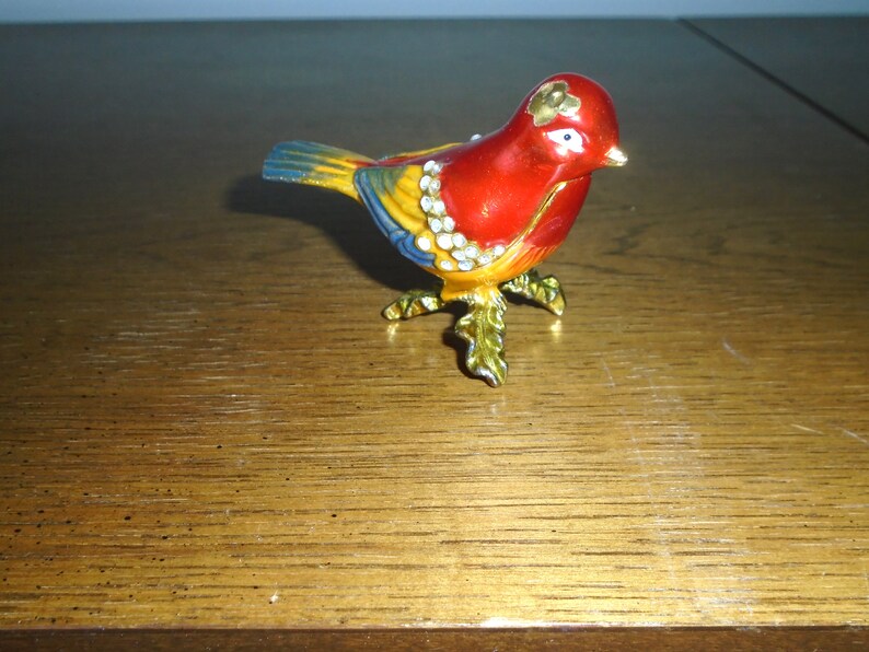 May include: A small, red, yellow, and blue bird figurine with gold accents and white rhinestones. The bird is perched on gold-colored legs with green leaves.
