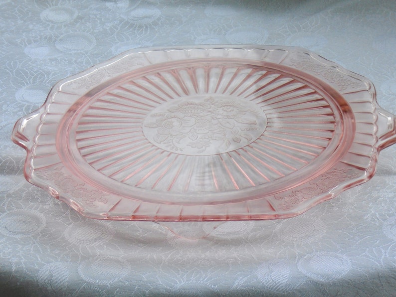 Puede incluir: Una bandeja de servir de cristal rosa con un dise&ntilde;o floral en el centro. La bandeja tiene forma rectangular con bordes festoneados y asas a cada lado. La bandeja es de cristal transparente y tiene un aspecto delicado y vintage.