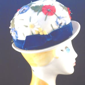 Puede incluir: Un sombrero blanco en forma de cúpula con un patrón floral vibrante de flores rojas, azules y blancas con hojas verdes. El sombrero está acentuado con una banda de cinta de terciopelo azul oscuro atada en un pequeño lazo en el lateral.