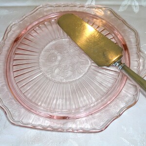 Puede incluir: Una fuente de pastel de vidrio rosa con un borde festoneado y un dise&ntilde;o floral en el centro. Un servidor de pastel dorado descansa sobre el plato, listo para servir un delicioso postre.