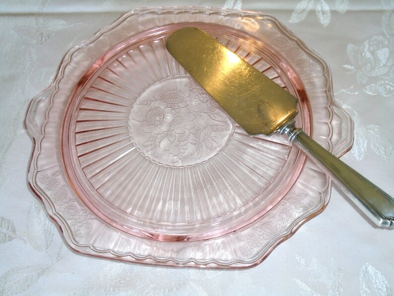 Puede incluir: Una fuente de pastel de vidrio rosa con un borde festoneado y un dise&ntilde;o floral en el centro. Un servidor de pastel dorado descansa sobre el plato, listo para servir un delicioso postre.