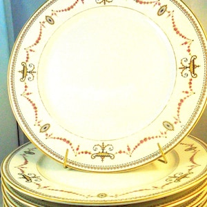 Puede incluir: Una pila de platos de porcelana color crema con bordes dorados y bordes decorativos. Los platos presentan un patrón repetitivo de guirnaldas florales rosas y detalles dorados. Los platos están apilados en un soporte dorado.