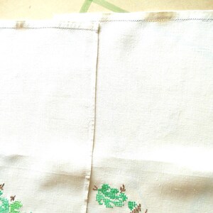 Puede incluir: Toalla de lino color crema con bordado floral de punto de cruz verde y marr&oacute;n a lo largo del borde inferior. La toalla est&aacute; dividida en dos paneles con una costura vertical. El borde superior tiene un dobladillo cosido.