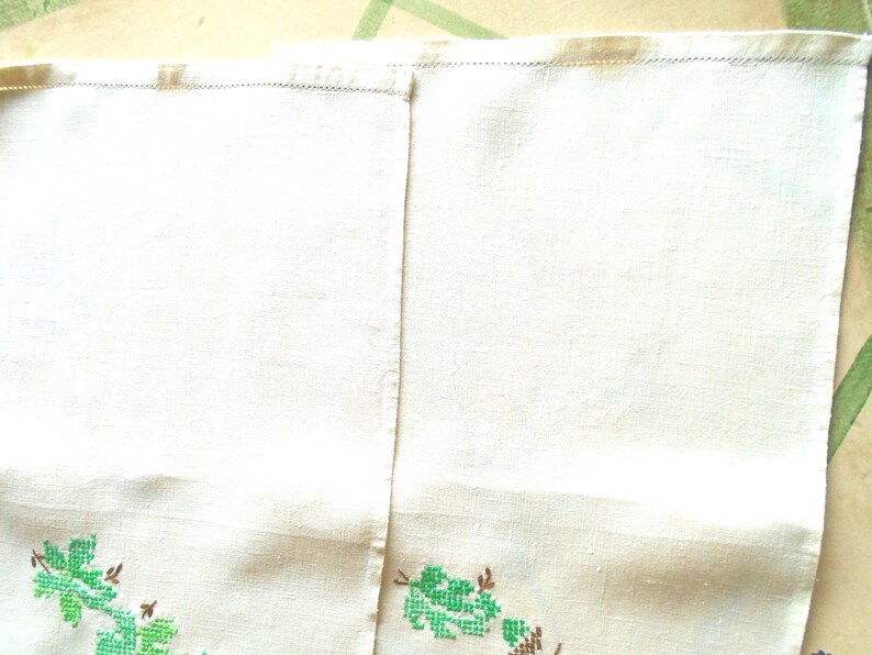 Puede incluir: Toalla de lino color crema con bordado floral de punto de cruz verde y marr&oacute;n a lo largo del borde inferior. La toalla est&aacute; dividida en dos paneles con una costura vertical. El borde superior tiene un dobladillo cosido.