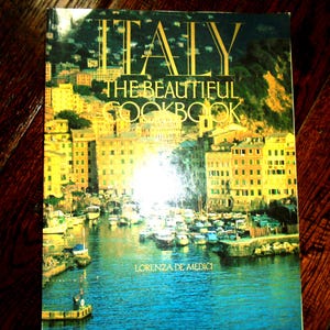 Puede incluir: Un libro de cocina de tapa dura titulado Italy The Beautiful Cookbook de Lorenza De Medici. La portada presenta una fotografía vibrante de una ciudad costera italiana con edificios amarillos, un puerto lleno de barcos y agua azul.