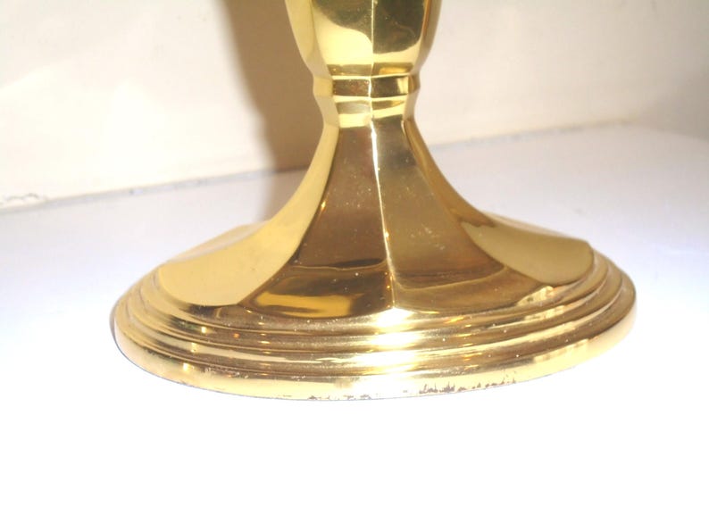 Puede incluir: Un candelero dorado con una base ancha y escalonada y un tallo acanalado. La base tiene una serie de anillos conc&eacute;ntricos y el tallo tiene un dise&ntilde;o facetado. El candelero est&aacute; hecho de un material brillante y reflectante.