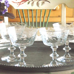 May include: Four clear champagne coupe glasses with etched floral patterns are displayed on a silver tray. The glasses have a classic shape and are arranged in a row. The background includes a chair and a potted plant.