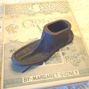 Puede incluir: Una horma de zapato de hierro fundido oscuro que descansa sobre un libro de tapa dura antiguo titulado The Golden West de Margaret Sidney. El objeto de metal presenta una punta redondeada y lisa y una sección de tobillo abierta con base ovalada.