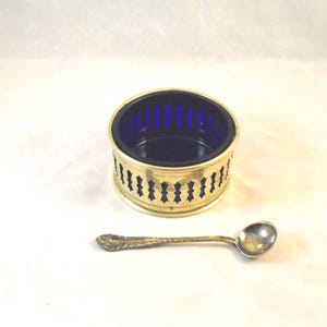 Puede incluir: Un salero vintage con exterior de metal dorado e interior de cristal azul cobalto. El salero tiene un diseño calado decorativo. Una pequeña cuchara ornamentada descansa junto a él.
