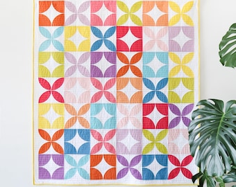 May include: A vibrant quilt featuring a repeating floral pattern in shades of orange, yellow, pink, purple, and blue against a white backdrop. The quilt has a yellow border and is displayed on a white wall. The quilt's design is reminiscent of a flower.