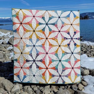 May include: A colorful quilt with a floral pattern. The quilt features a repeating design of white petals with accents of pink, orange, yellow, blue, and navy. The quilt is displayed outdoors against a backdrop of a lake and rocky shoreline.