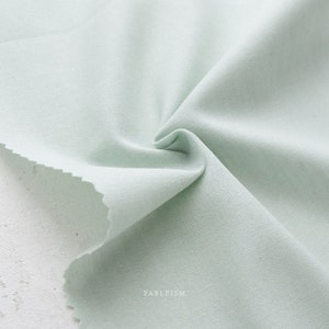 May include: A close-up of a light green fabric with a smooth texture. The fabric is folded and draped, showing its soft and drapey quality.