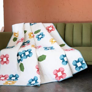 May include: A white quilt with a floral pattern in shades of pink, blue, green, and yellow. The quilt is folded and draped over a green sofa.