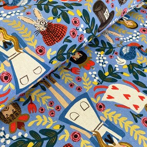 May include: Blue fabric with a whimsical pattern featuring Alice in Wonderland characters, including Alice, the Mad Hatter, the White Rabbit, and a Cheshire Cat. The fabric is filled with colourful flowers, leaves, and other details.