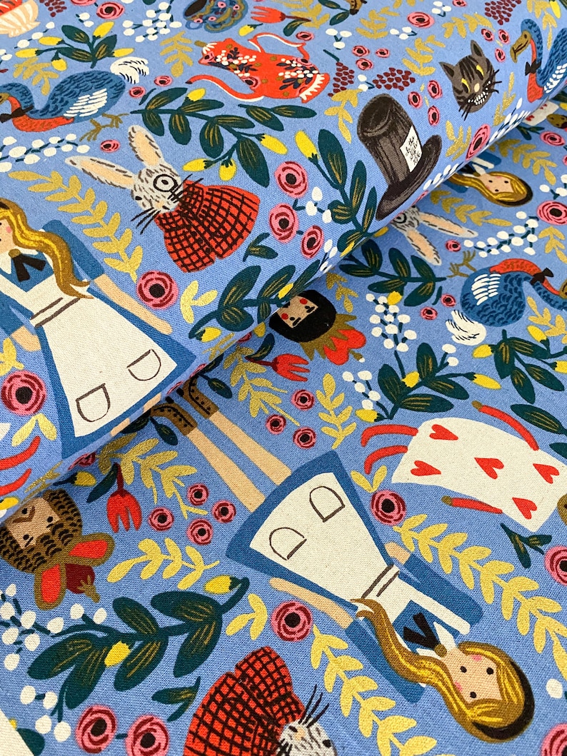 May include: Blue fabric with a whimsical pattern featuring Alice in Wonderland characters, including Alice, the Mad Hatter, the White Rabbit, and a Cheshire Cat. The fabric is filled with colourful flowers, leaves, and other details.