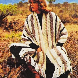 May include: A striped crocheted shawl with fringe, in shades of white, brown, and black. The shawl is draped over a person's shoulders, and the person is sitting on a rock.
