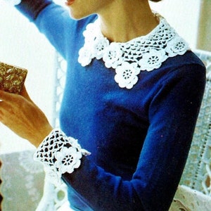 Op de afbeelding: Een vrouw draagt een blauwe trui met een witte gehaakte kraag en manchetten. Ze houdt een klein, versierd, gouden doosje vast. De trui heeft een witte gehaakte kraag en manchetten.