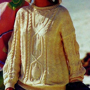 May include: A cream-colored knitted sweater with a cable knit design. The sweater has a round neckline and long sleeves.