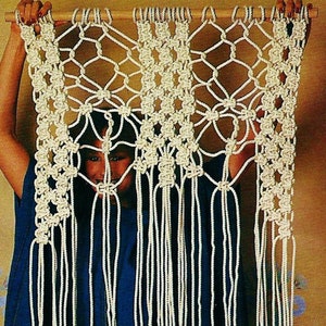 May include: A white macrame wall hanging with a geometric pattern. The hanging is made of knotted cords and has a wooden dowel at the top.
