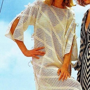 Puede incluir: Un vestido blanco de crochet de corte holgado y estampado geométrico. El vestido tiene mangas largas con aberturas en las muñecas y lazos en los puños. El vestido se muestra en una modelo de pie contra un fondo de cielo azul.