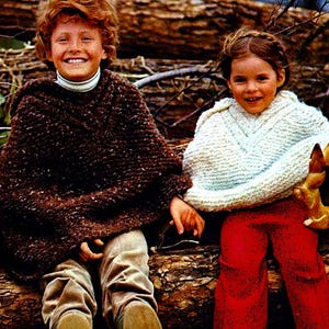 May include: Two children wearing knitted ponchos, one in brown and one in white, are sitting on a log. The child in the brown poncho is smiling and has red hair. The child in the white poncho has long brown hair and is wearing red trousers.