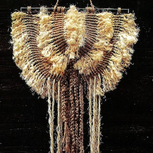 May include: A brown and beige macrame wall hanging with a fringe design. The hanging is made of natural fibers and has a rustic, bohemian style.