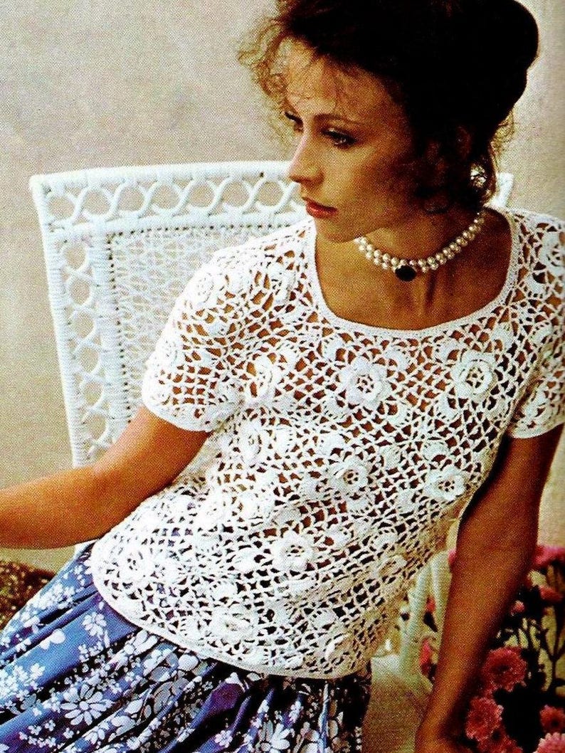 May include: A white crochet top with a delicate floral pattern. The top has a scoop neckline and short sleeves. The model is wearing a blue floral skirt.