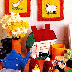 Könnte beinhalten: Ein Set gestrickter Küchenaccessoires mit einem hausförmigen Teekannenwärmer, zwei Eierwärmern und zwei Topflappen. Die Topflappen zeigen eine Ente und ein Schaf. Die Eierwärmer sind rot und weiß mit einem Enten- und Schafdesign. Der Teekannenwärmer ist rot, weiß und grün mit einem Hausdesign.