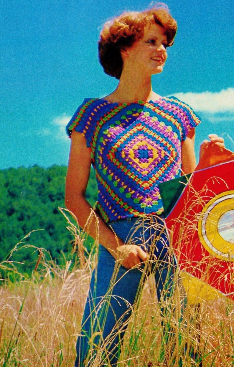 Puede incluir: Una mujer lleva una blusa de ganchillo de colores con un patr&oacute;n cuadrado en tonos de azul, morado, amarillo y verde. Est&aacute; de pie en un campo de hierba alta y sostiene una cometa roja, amarilla y verde.