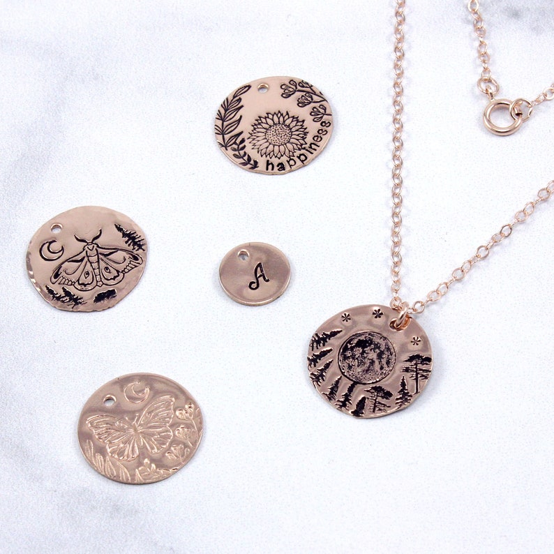 K&ouml;nnte beinhalten: Ein Set aus f&uuml;nf ros&eacute;goldfarbenen Metall-Anh&auml;ngern mit verschiedenen gravierten Designs. Die Anh&auml;nger umfassen einen Schmetterling, eine Motte, eine Sonnenblume mit dem Wort "happiness" darum herum, einen Vollmond mit B&auml;umen und einen Anh&auml;nger mit dem Buchstaben "A".