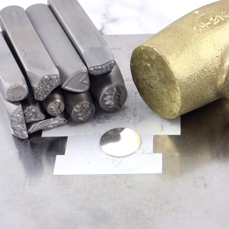 K&ouml;nnte beinhalten: Ein Satz Metallstempel mit verschiedenen Designs, darunter ein Herz, eine Blume und ein Blatt. Die Stempel sind auf einer Metalloberfl&auml;che neben einem Messinghammer angeordnet.