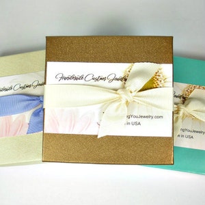 May include: Three gift boxes with gold, beige and teal lids. The gold box has a white ribbon and a label that reads "Handmade Custom Jewelry igYouJewelry.com in USA".