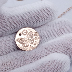 K&ouml;nnte beinhalten: Eine goldfarbene Halskette mit einem runden Anh&auml;nger, der ein filigranes Design aus einem Schmetterling, einem Halbmond und floralen Elementen zeigt.