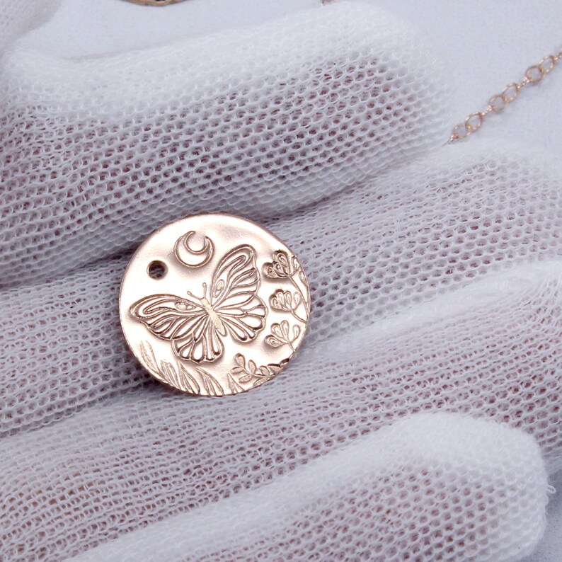 K&ouml;nnte beinhalten: Eine goldfarbene Halskette mit einem runden Anh&auml;nger, der ein filigranes Design aus einem Schmetterling, einem Halbmond und floralen Elementen zeigt.