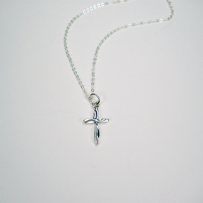May include: A silver chain necklace with a small, silver cross pendant. The cross is a simple design with a twisted or knotted look.