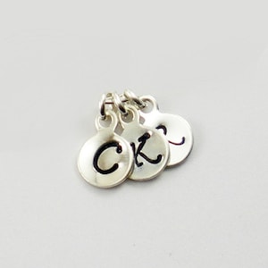 May include: Three small, silver-colored circular charms with individual letters. The charms are connected by small loops. The letters "C", "K", and "?" are visible on the charms. The background is a solid, light gray.