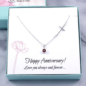 May include: A silver necklace with a cross design and a small, round, red gemstone pendant. The necklace is presented in a light blue gift box with the words "Happy Anniversary! Love you always and forever..."