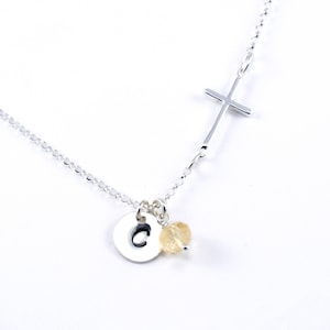 May include: Silver necklace with a cross charm, a small round charm with the letter C, and a light yellow bead.