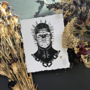 May include: A black and white illustration of a figure with pins in their head and a chain-like design on the collar. The artwork is on a textured, off-white paper. Dried flowers and foliage surround the artwork.