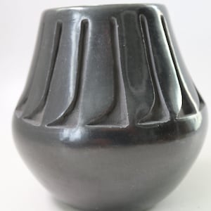 May include: A small, rounded black vase with a smooth, polished finish. The upper portion of the vessel features a series of vertical, carved indentations that create a rhythmic, textured pattern around the circumference of the neck.